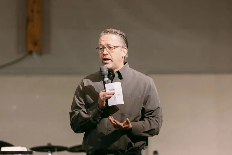 A man wearing glasses and a gray shirt speaking into a microphone while holding a note card, standing against a plain background.
