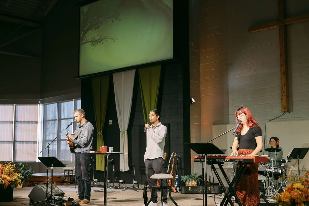 A band performing on stage with three musicians singing and playing instruments, including a guitar, keyboard, and drums, in a church or community hall setting with large windows and a cross on the wall.