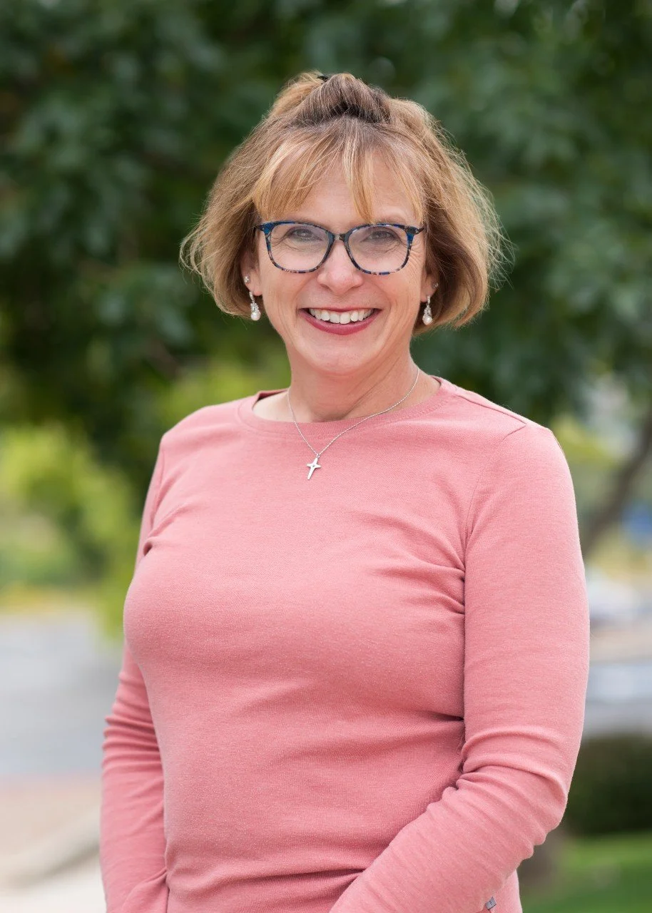 A smiling woman with short blonde hair, glasses, and pearl earrings wearing a pink long-sleeve top, standing outdoors with green trees in the background.