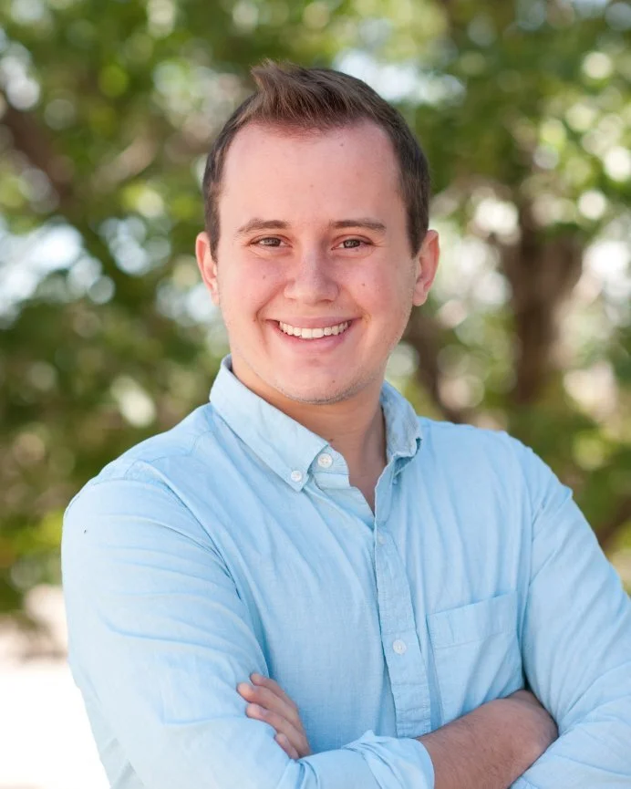 Smiling young man with short brown hair wearing a light blue button-up shirt, standing outdoors with green trees in the background.