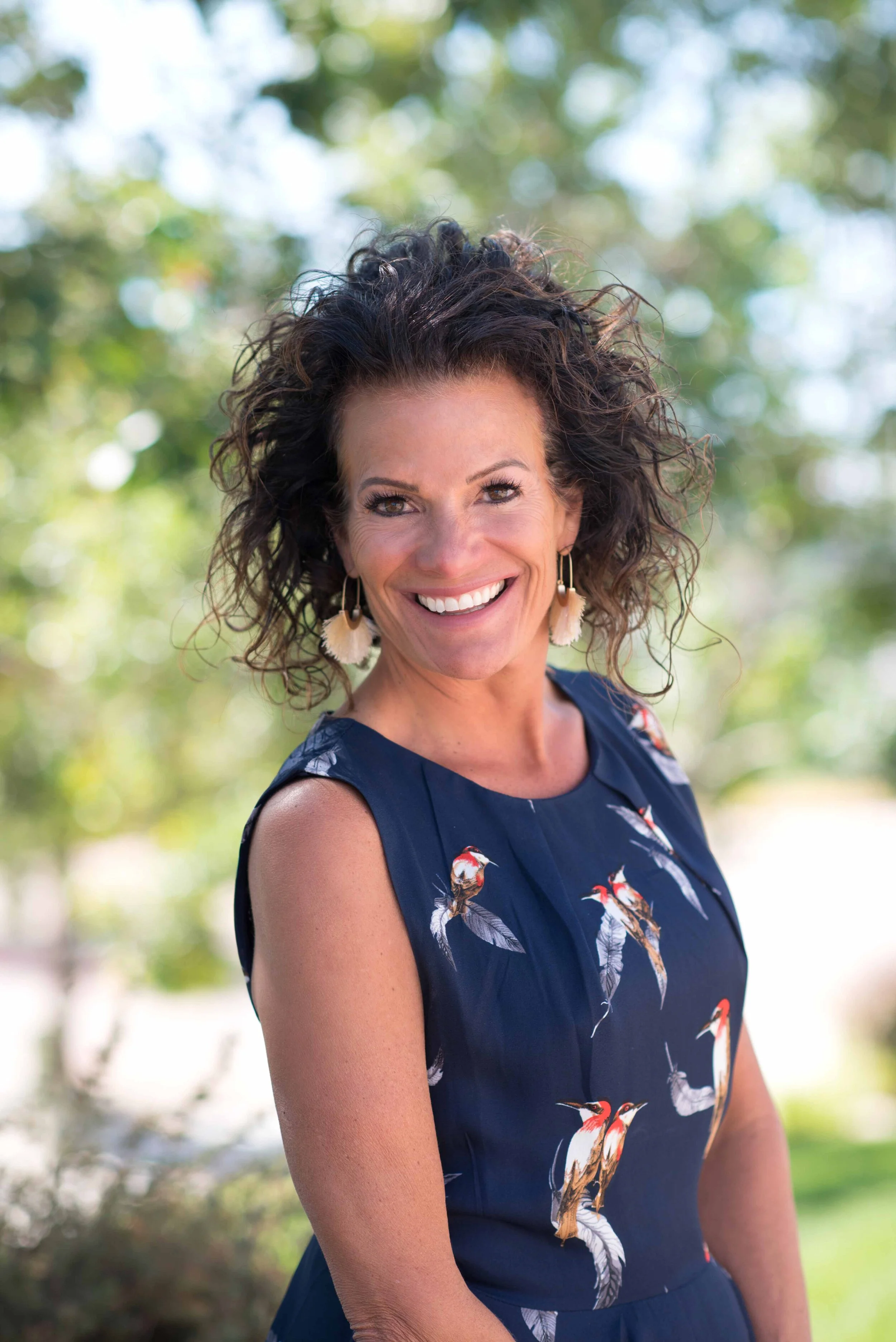 A smiling woman with curly dark hair, wearing a sleeveless navy blue dress with a bird pattern, earrings, and standing outdoors with a blurred background of trees and greenery.