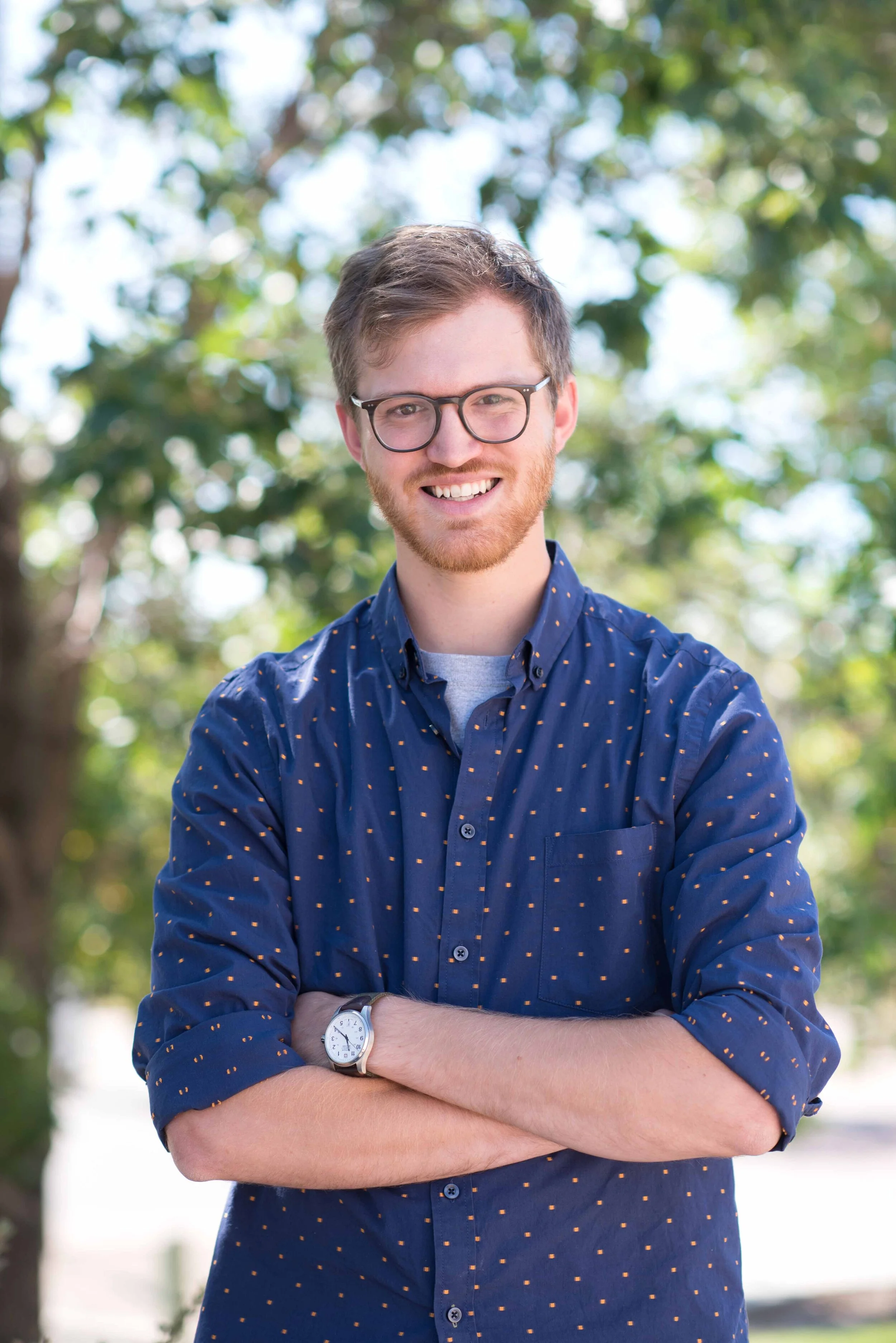 A young man with glasses and a beard, smiling with arms crossed, wearing a blue shirt with small yellow patterns, standing outdoors in front of green trees.
