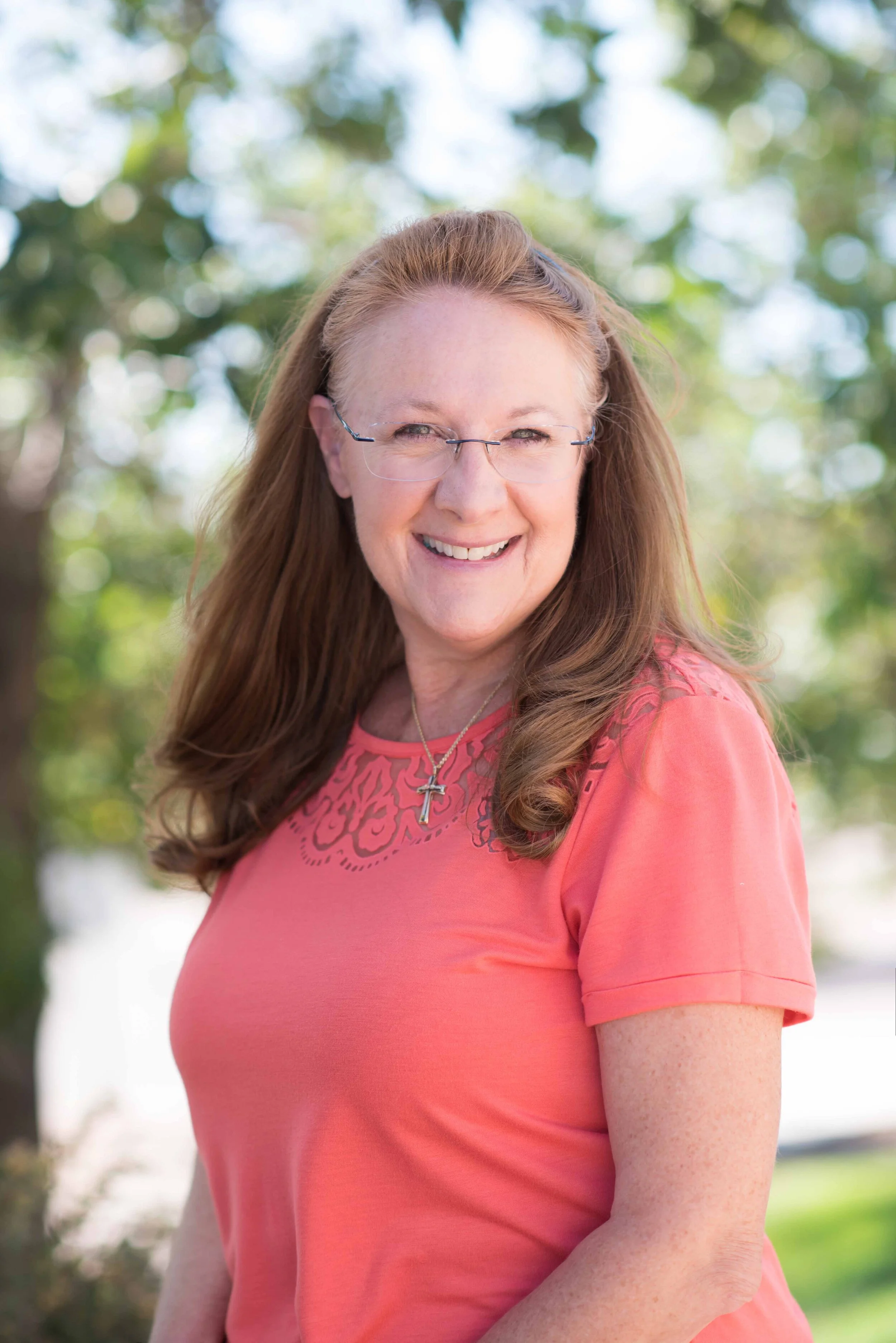 A cheerful woman with long red hair, wearing glasses, a peach-colored shirt with lace detail, and a necklace with a cross, standing outdoors with trees in the background.