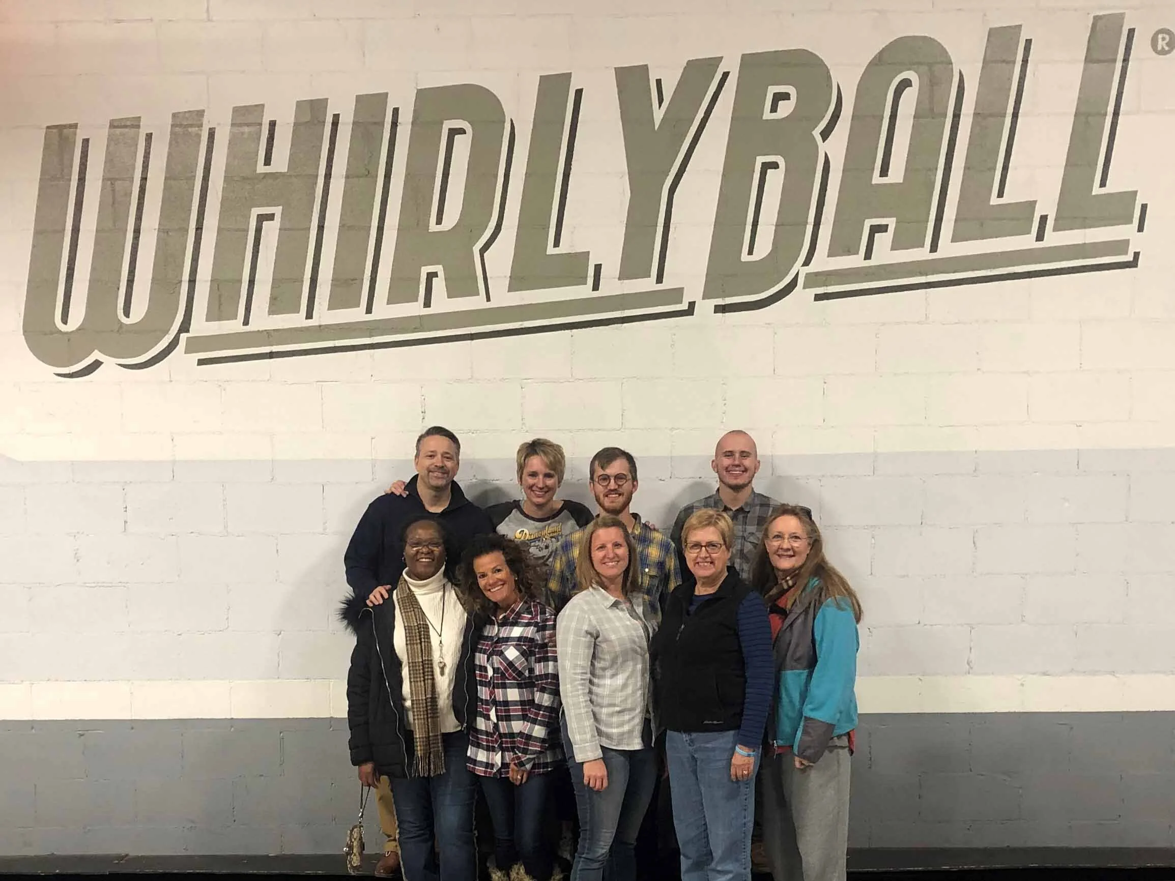 Group of nine smiling people standing in front of a large painted sign that says "WORLIDYALL" on a white brick wall.