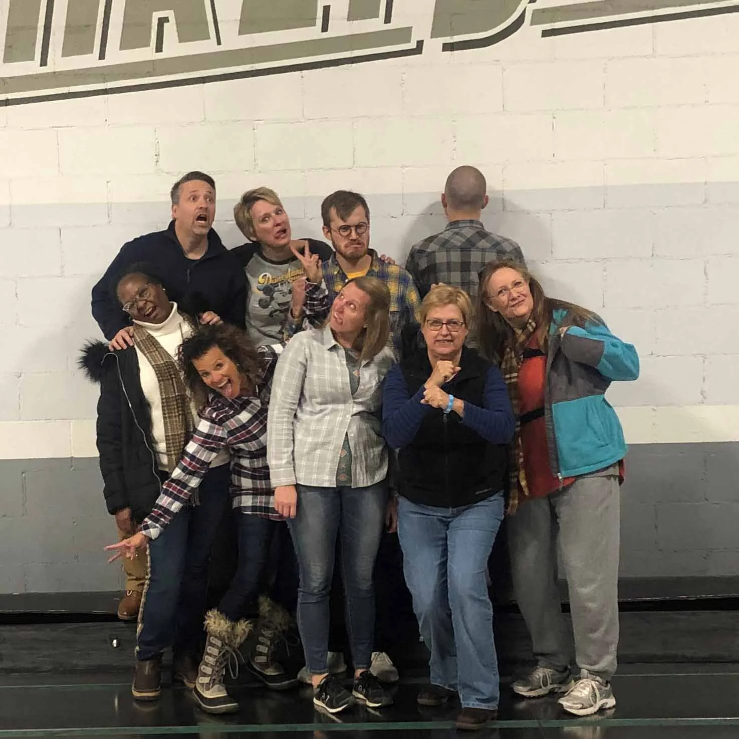 A group of ten people posing in front of a white brick wall, with some making funny faces and gestures.