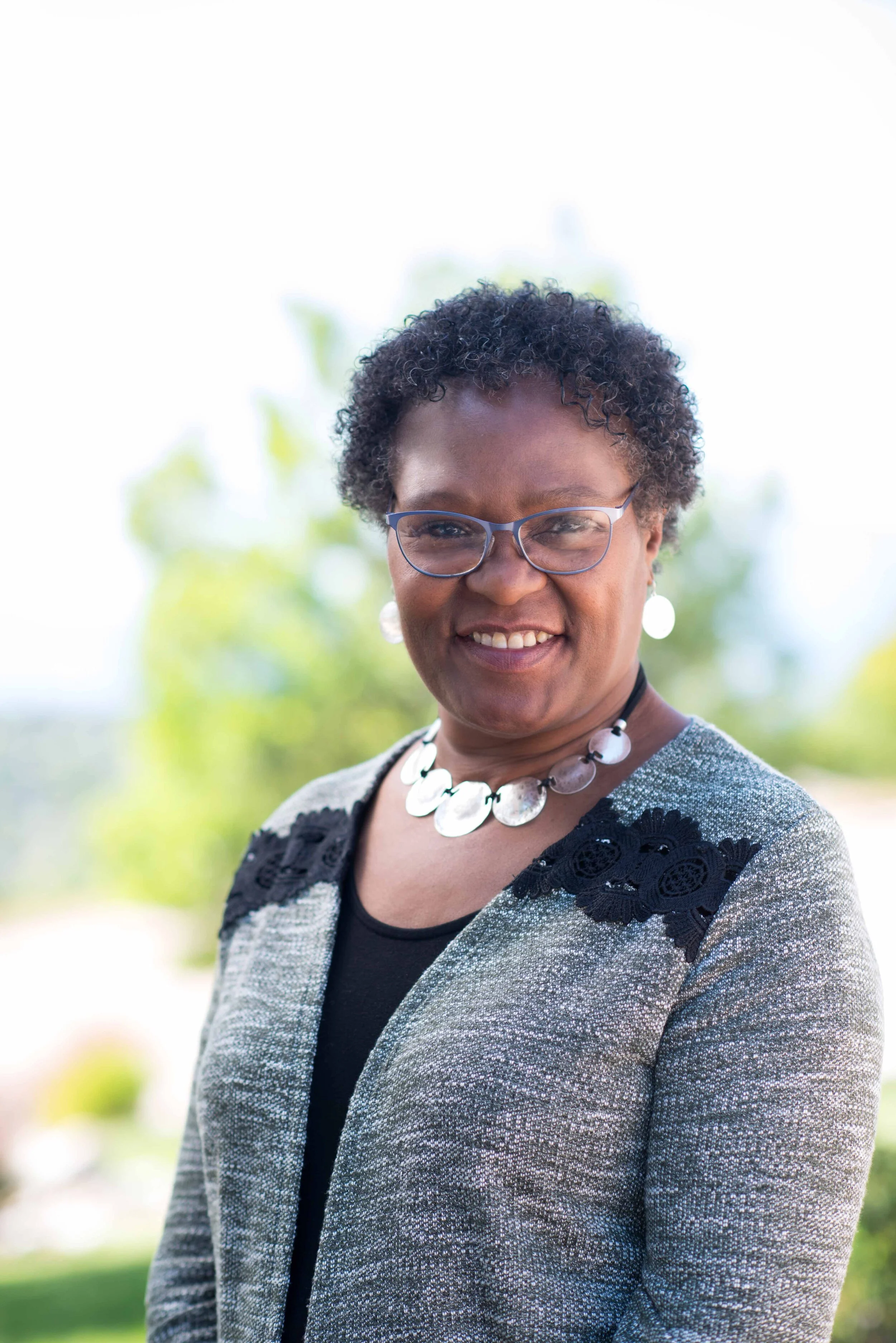 A middle-aged woman with short curly hair and glasses, smiling outdoors with a blurred green background, wearing a black top, a gray blazer with black lace details, a statement necklace, and matching earrings.