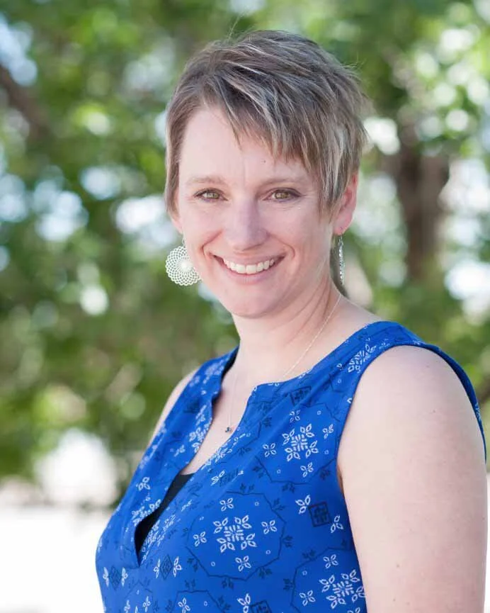 A woman with short brown hair wearing a sleeveless blue dress with white patterns, smiling outdoors with green trees in the background.