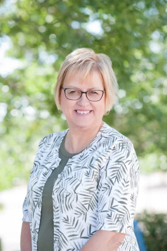 A smiling woman with short blond hair, glasses, and a patterned blouse standing outdoors in front of a blurred background of green trees.
