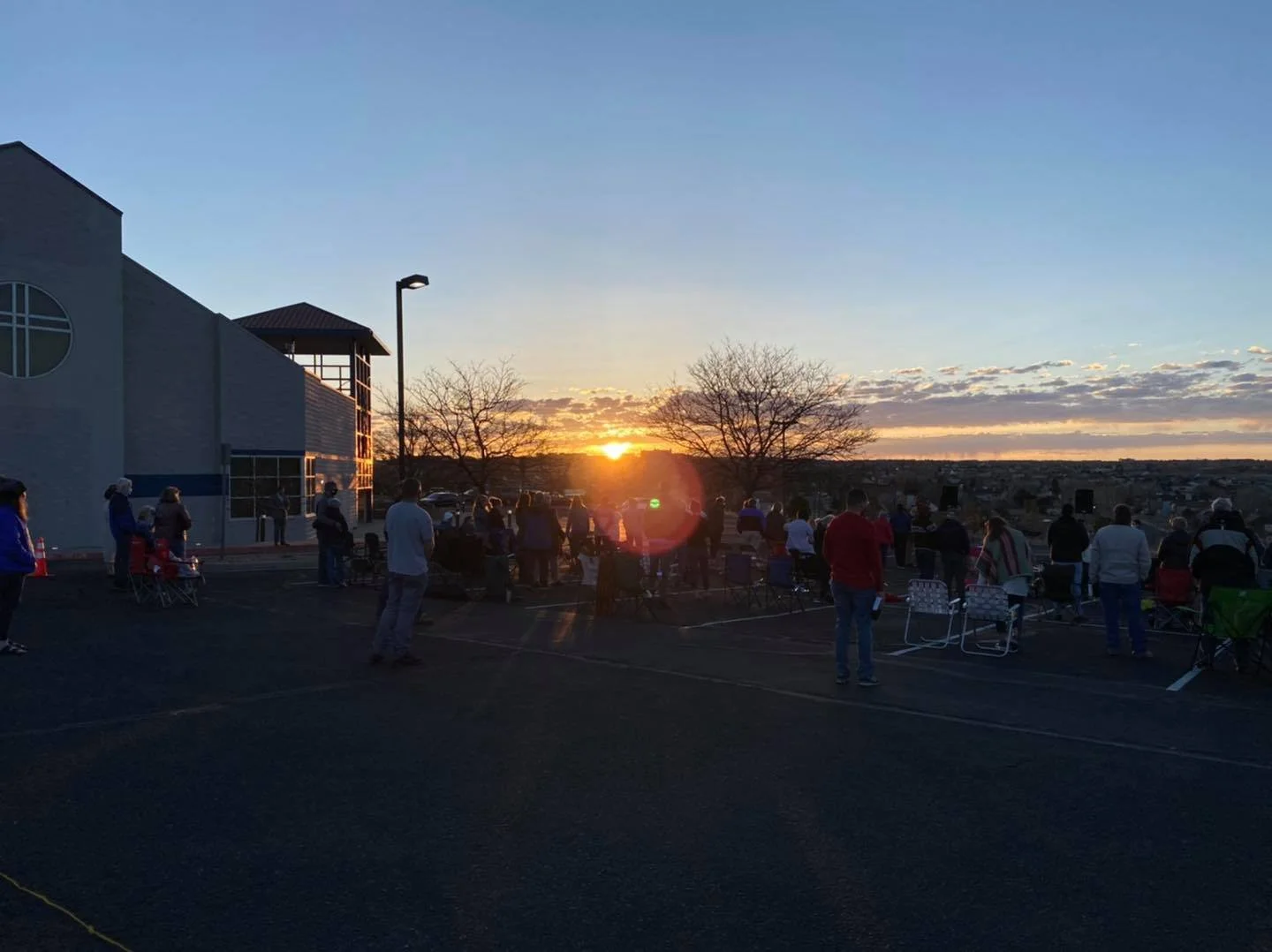 People gathered at an outdoor event during sunset, with some standing and sitting in chairs, near a building on the left, and a tree line in the distance.