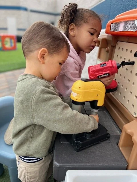 Two young children using toy power tools at a pretend workbench outdoors.