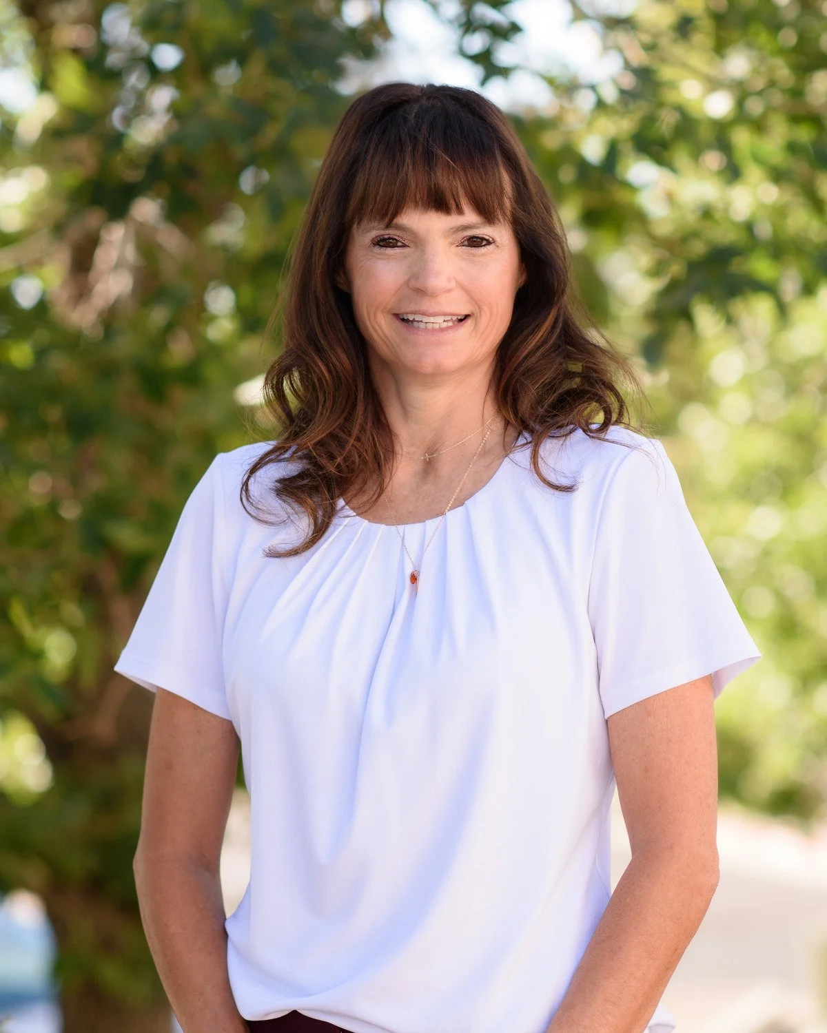 A woman with brown hair and bangs, wearing a white top and a gold necklace, smiling outdoors with green trees and sunlight in the background.