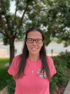 Woman with long dark hair wearing glasses and a pink t-shirt standing outdoors in front of trees.
