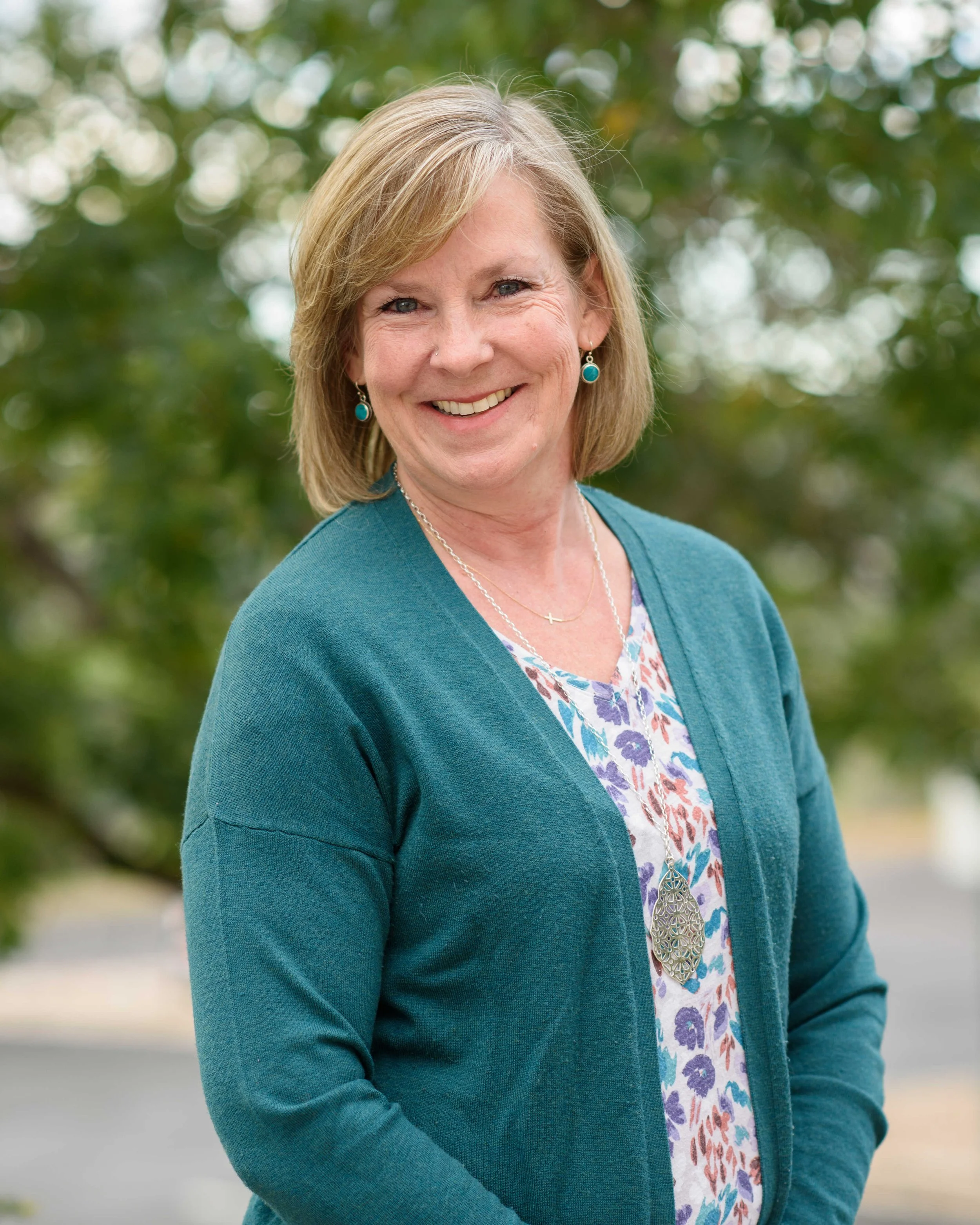 A woman with shoulder-length blonde hair smiling outdoors, wearing a teal cardigan, patterned blouse, turquoise earrings, and a necklace with a large pendant, with trees in the background.