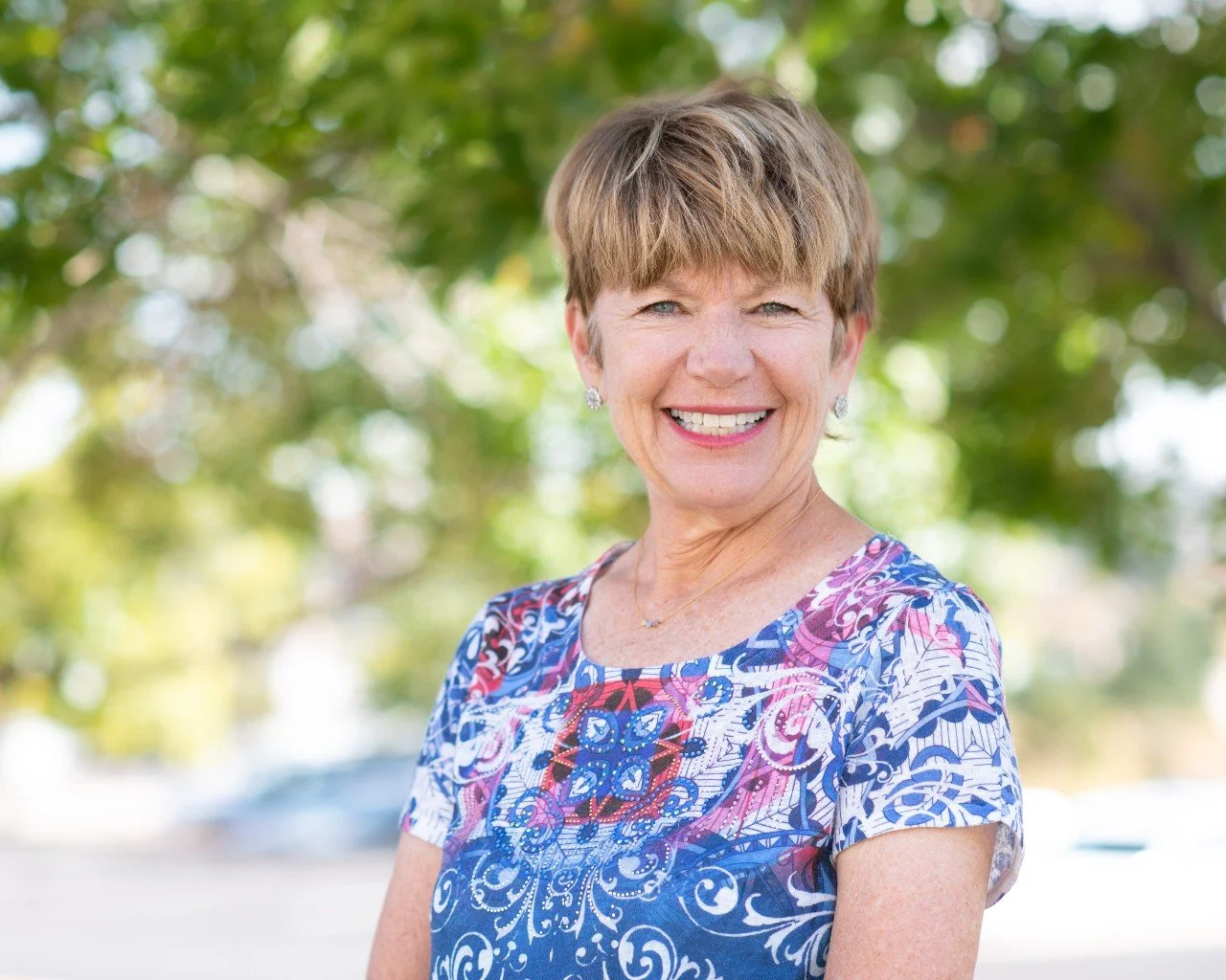 A smiling woman with short light brown hair wearing a colorful, patterned blouse standing outdoors with a blurred green tree background.