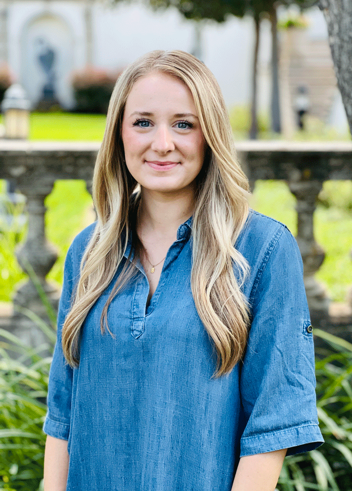 A young woman with long blonde hair and blue eyes, wearing a blue denim shirt, standing outdoors in a park-like setting with grass, trees, and stone structures in the background.