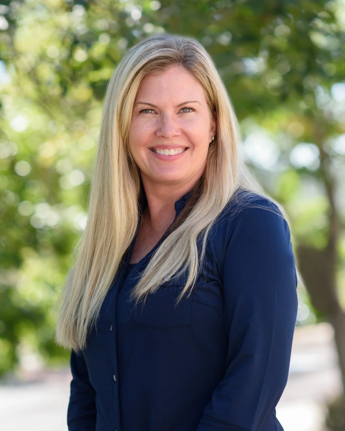 A woman with long blonde hair smiling outdoors in front of green trees, wearing a dark blue shirt.