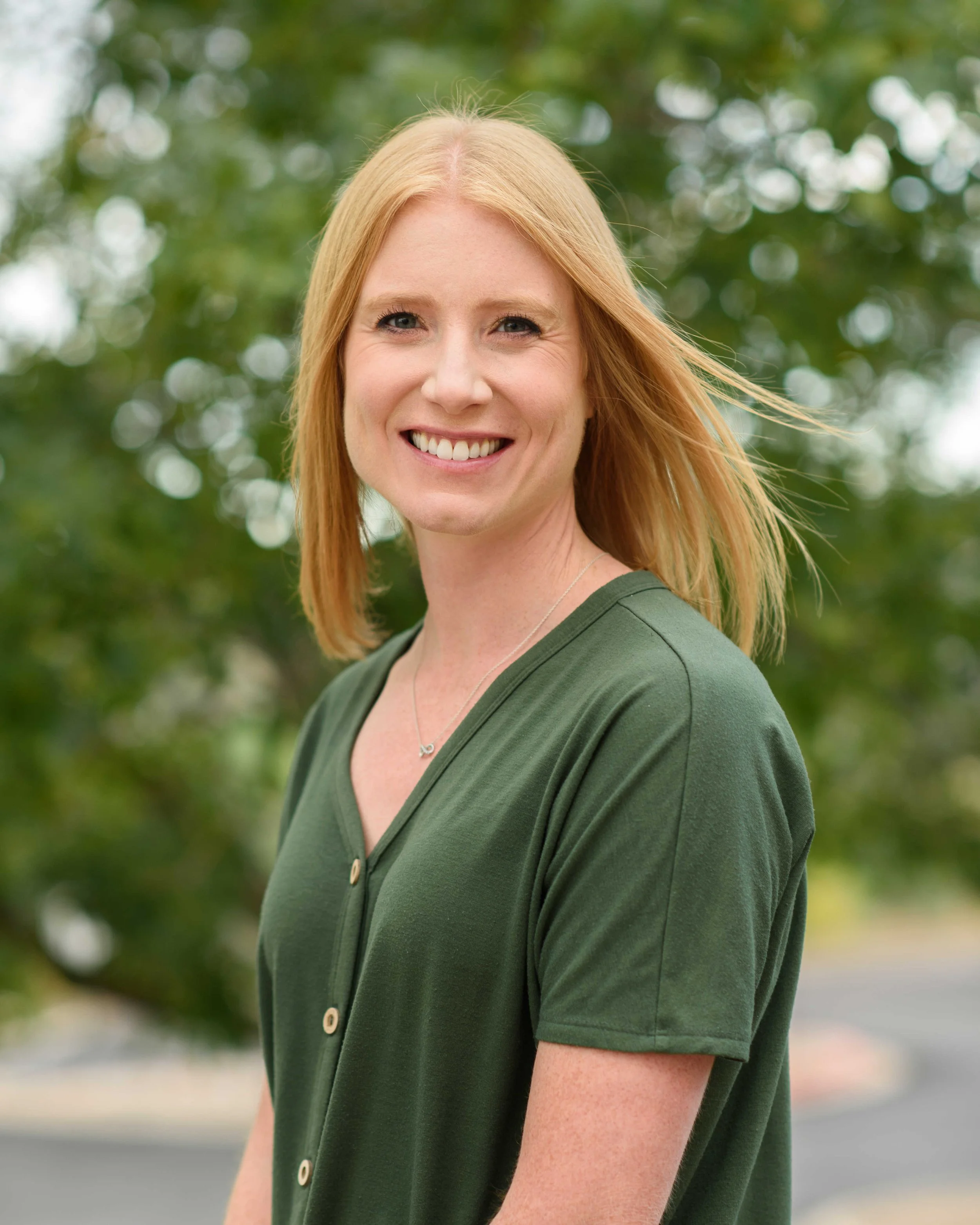 A woman with shoulder-length red hair smiling outdoors in front of green blurred trees, wearing a dark green shirt and a necklace.