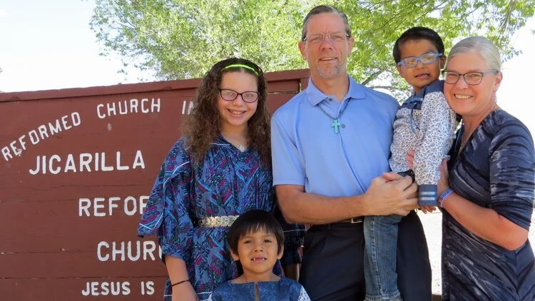 A family of five stands outdoors in front of a sign for Jicarilla Reformed Church, smiling and posing for a photo.