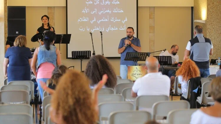 People attending a church service with a man speaking at the pulpit, a woman singing, and a keyboard player, with Arabic text projected on a screen behind them.