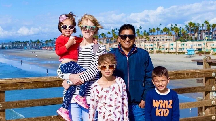 Family of five on a beach boardwalk, smiling, with houses and palm trees in the background.