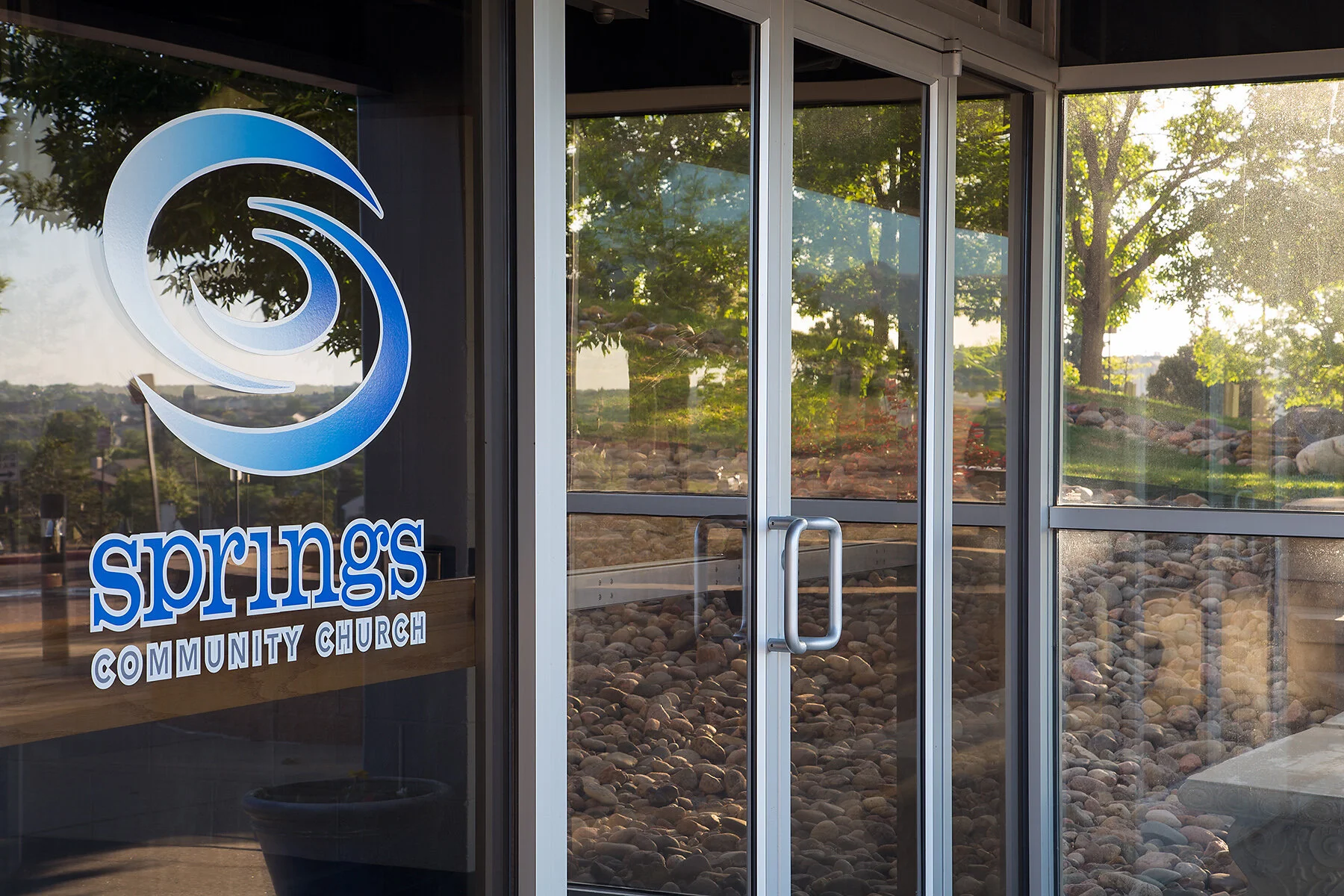 Glass door of Springs Community Church with a wooden bench inside and a large logo sign visible, showing the church name and a stylized wave symbol, with trees and sunlight outside.