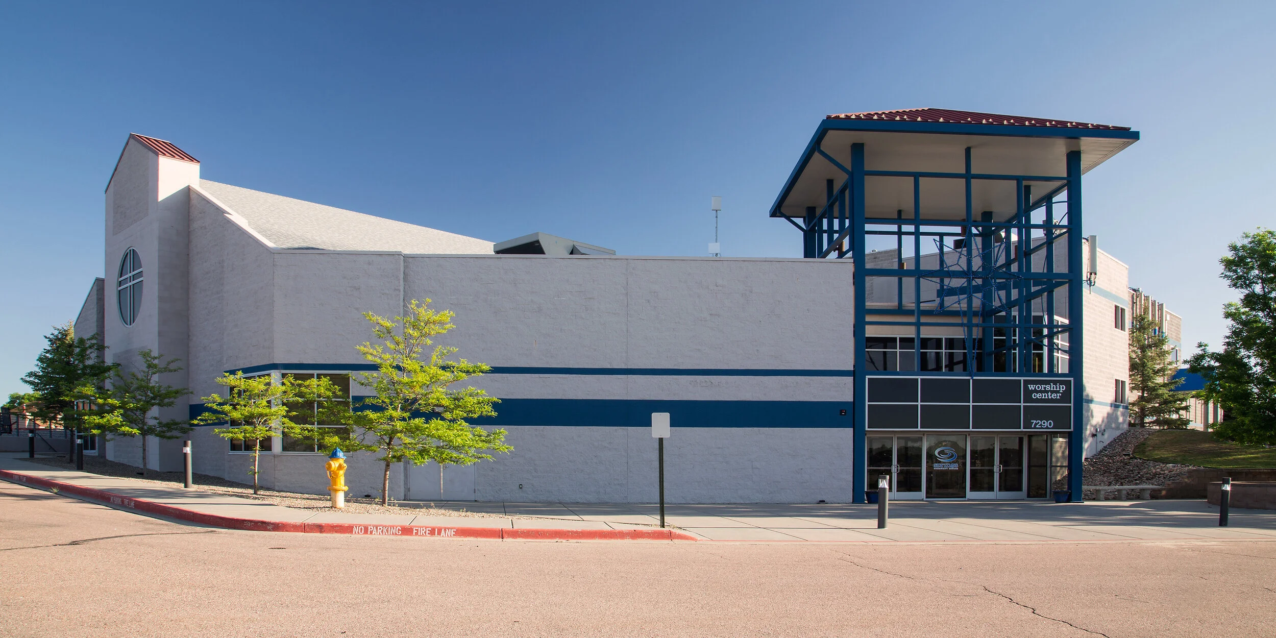 Exterior of a church or worship center with white walls, blue accents, a stairway, and trees in front. Sign reads 'worship center' and the address is 7290.