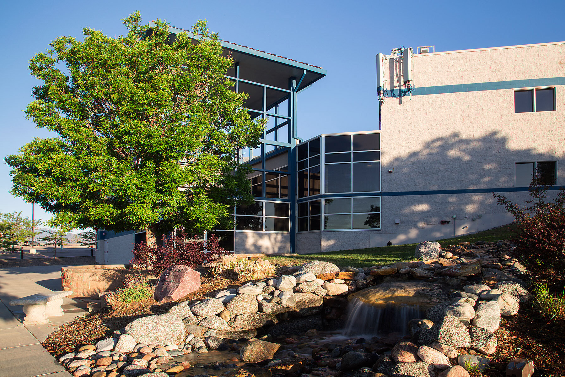 Modern building with large glass windows, landscaping with rocks, a small waterfall, and trees, under a clear blue sky.