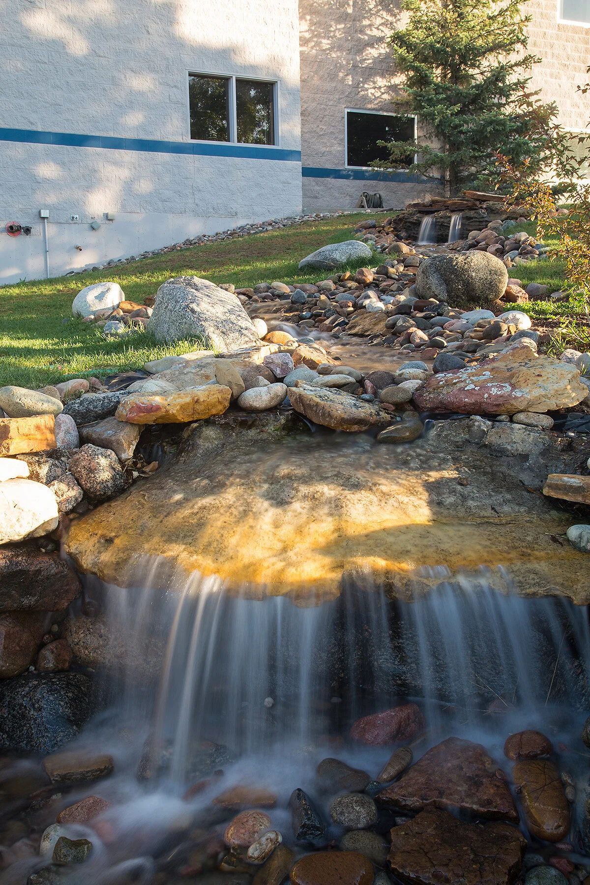 Small waterfall flowing over rocks in a landscaped outdoor area with grass, trees, and a building in the background.