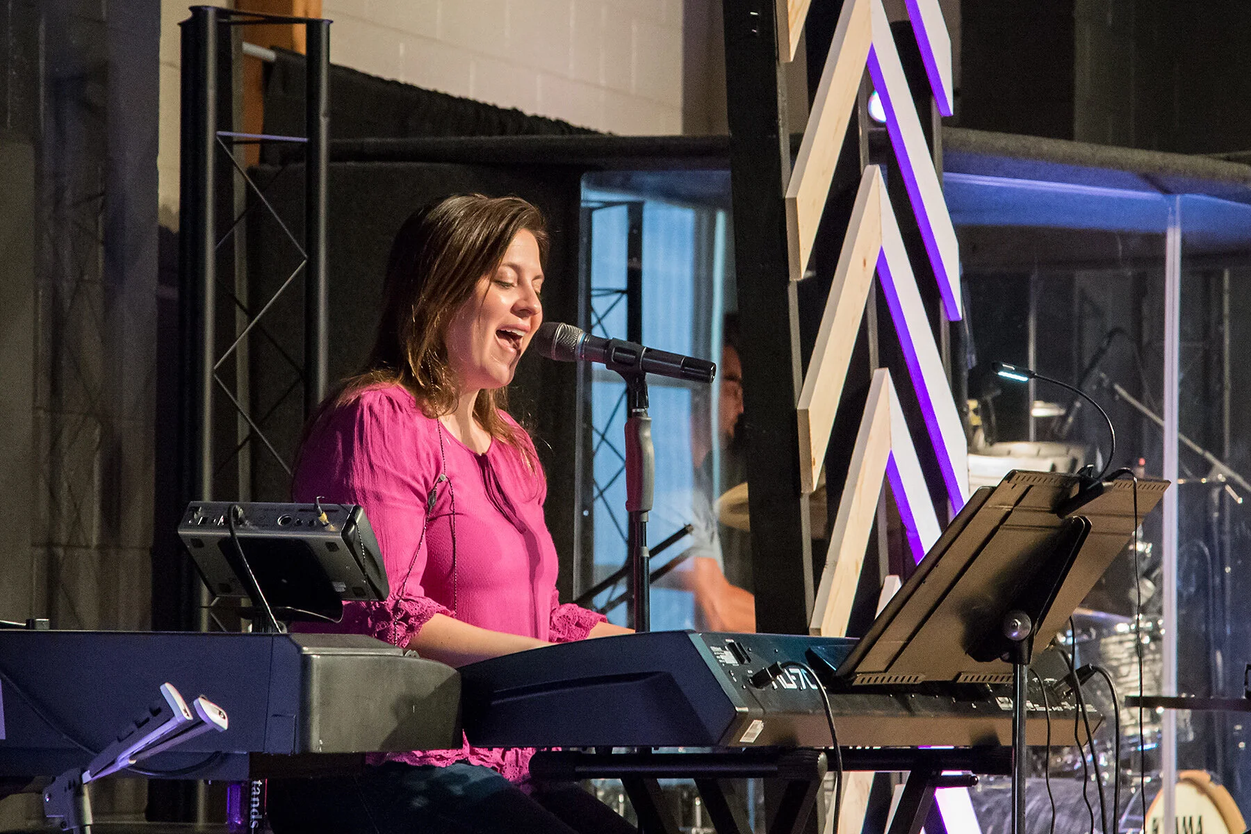 A woman singing and playing keyboard on stage with a microphone in front of her, with a man in the background and stage lighting.