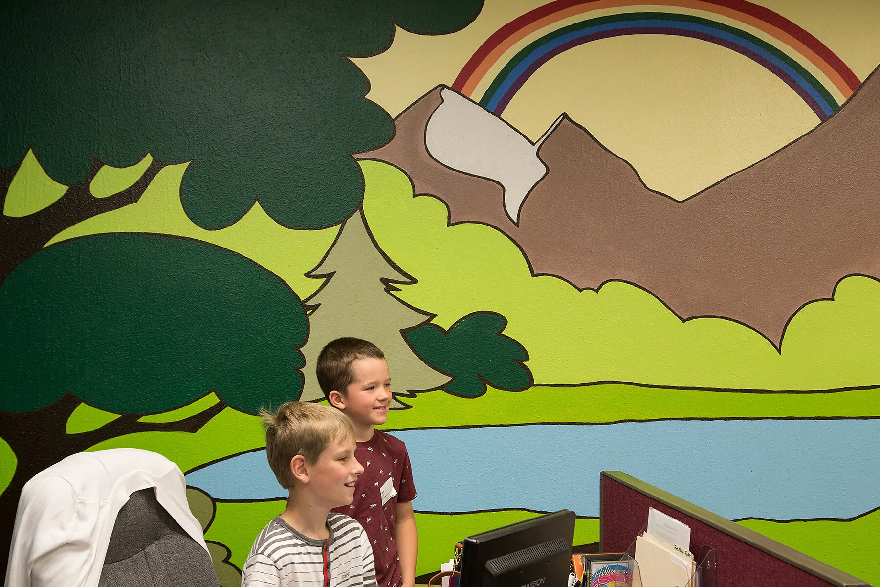 Two young boys standing in front of a colorful mountain and river mural at an indoor location.