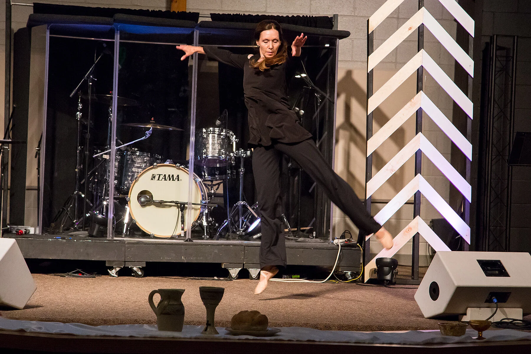 A woman in black performing a dance on a stage with musical instruments, including a drum set behind her, and decorative elements, such as a zigzag patterned backdrop.