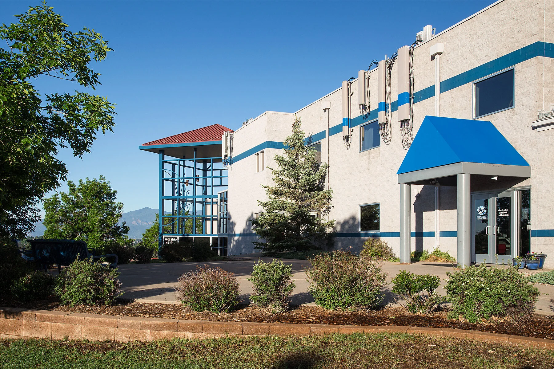Modern building with a blue roofed entrance and a sign reading worship center, surrounded by bushes and trees, with mountains in the background under a clear blue sky.