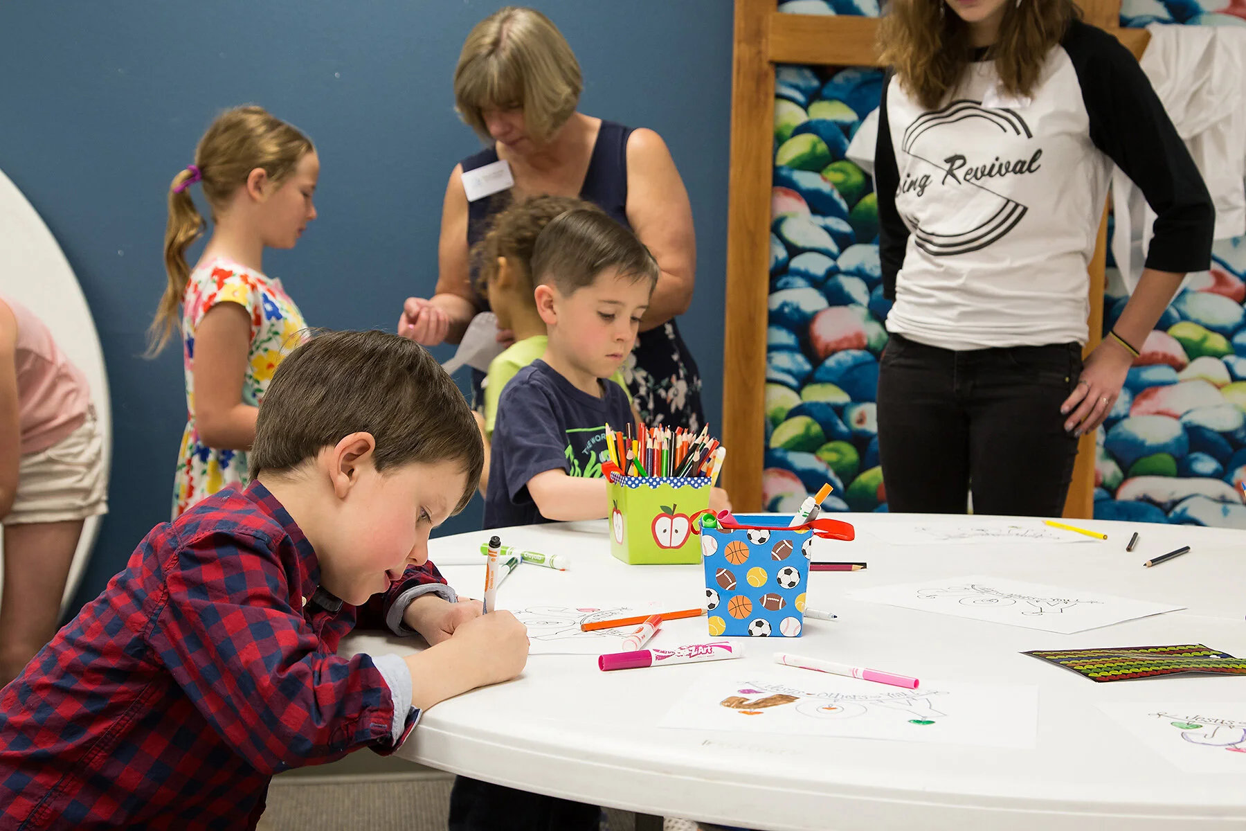 Children sitting and standing around a table engaged in coloring activities, with markers and drawings, in a colorful indoor setting.