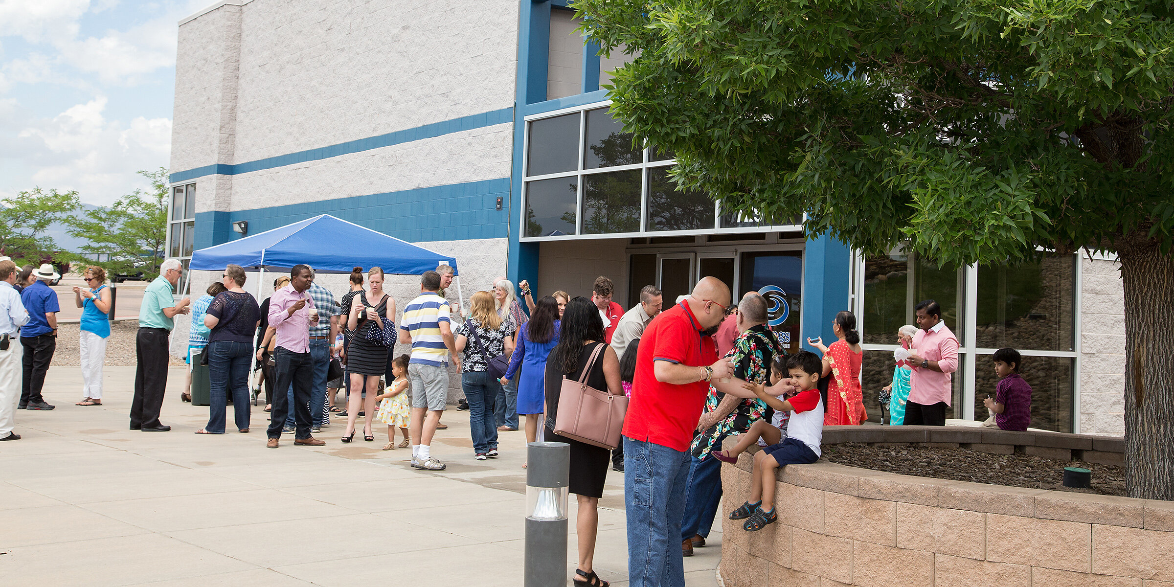 People standing in line outside a building, some using their phones, under a blue canopy, with a large tree on the right side, and a glass entrance door.