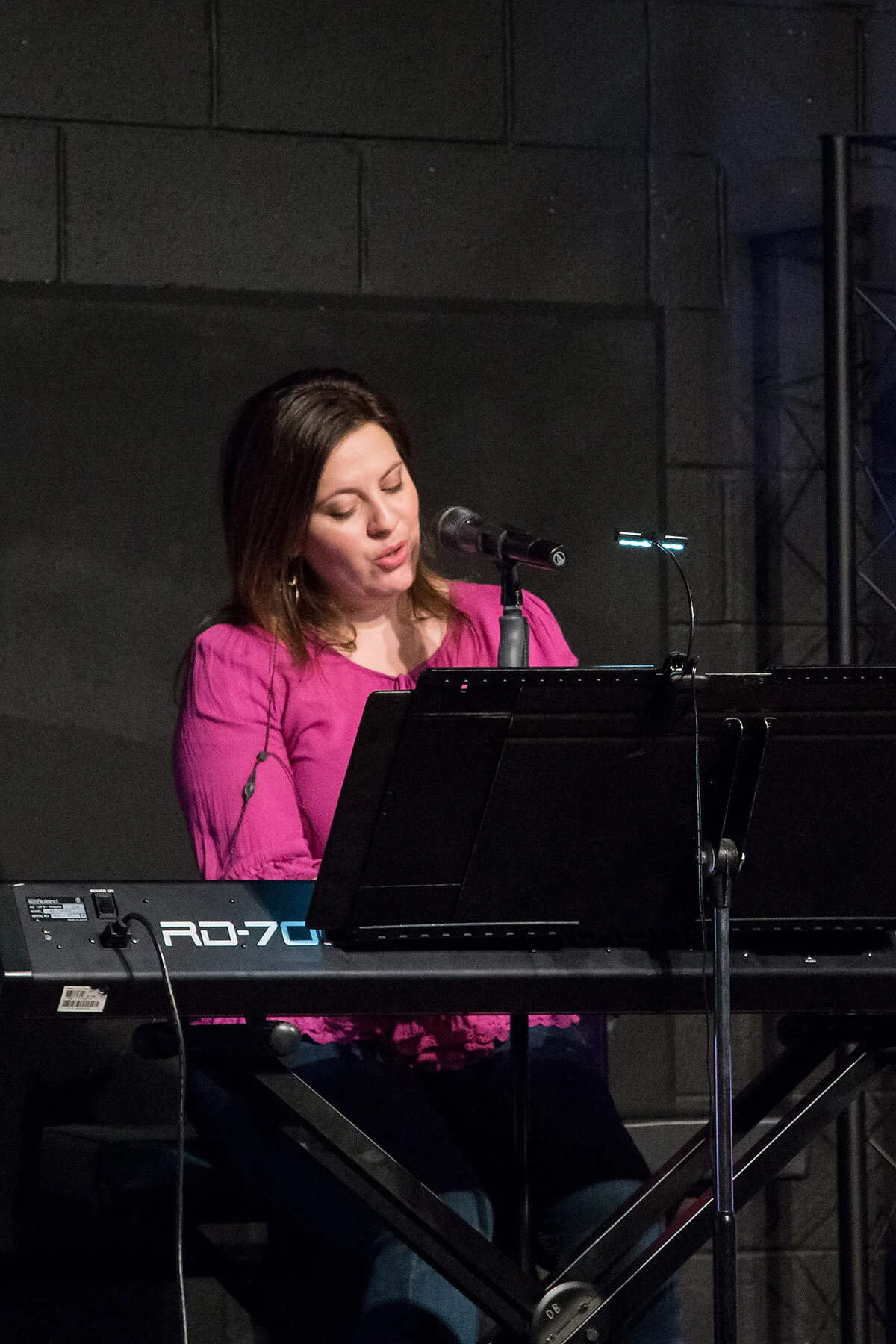 A woman singing or speaking into a microphone while playing a keyboard on stage.