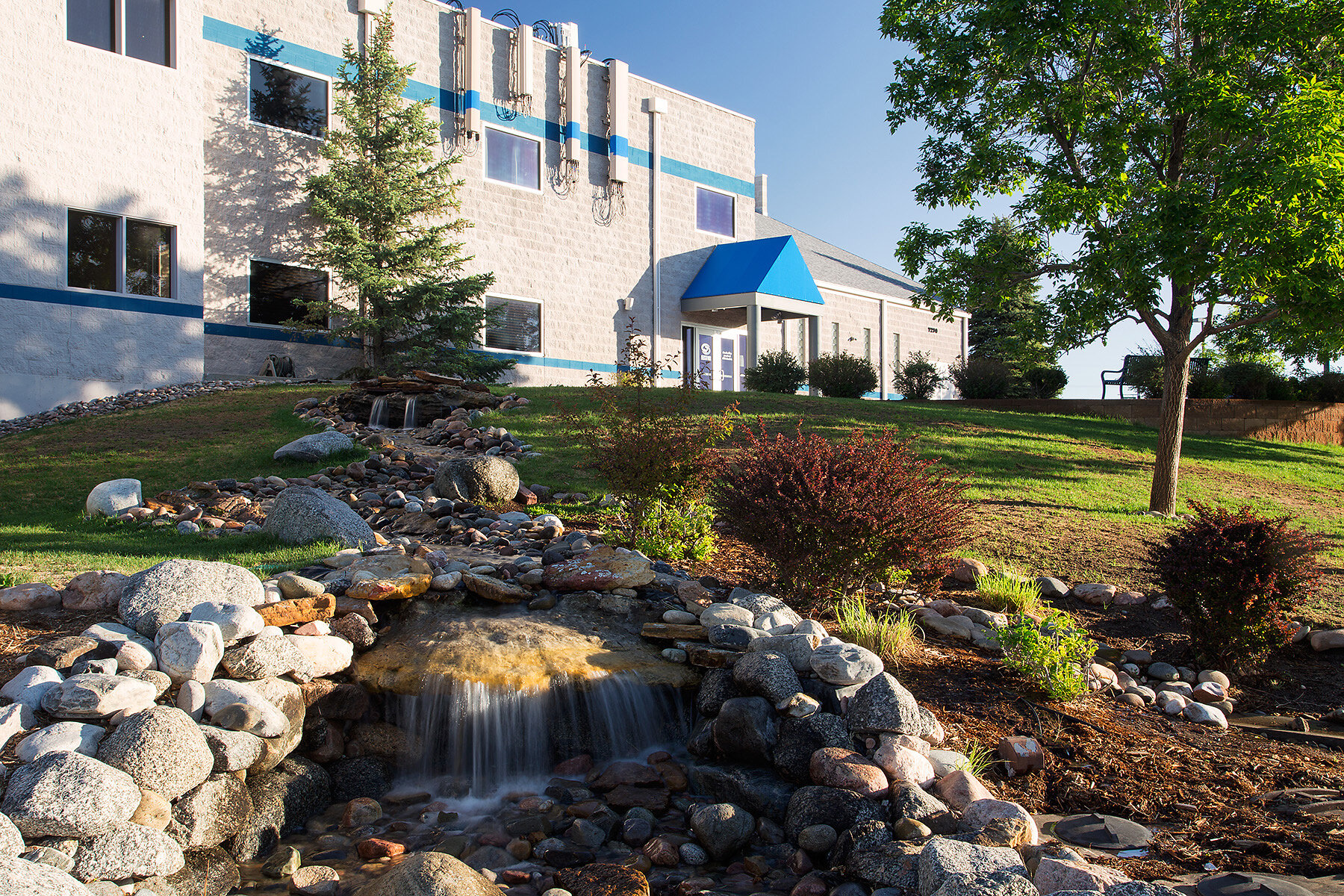 A landscaped view of a building with a blue awning, a small waterfall feature with rocks and stones, and green trees and bushes in the foreground and background on a sunny day.