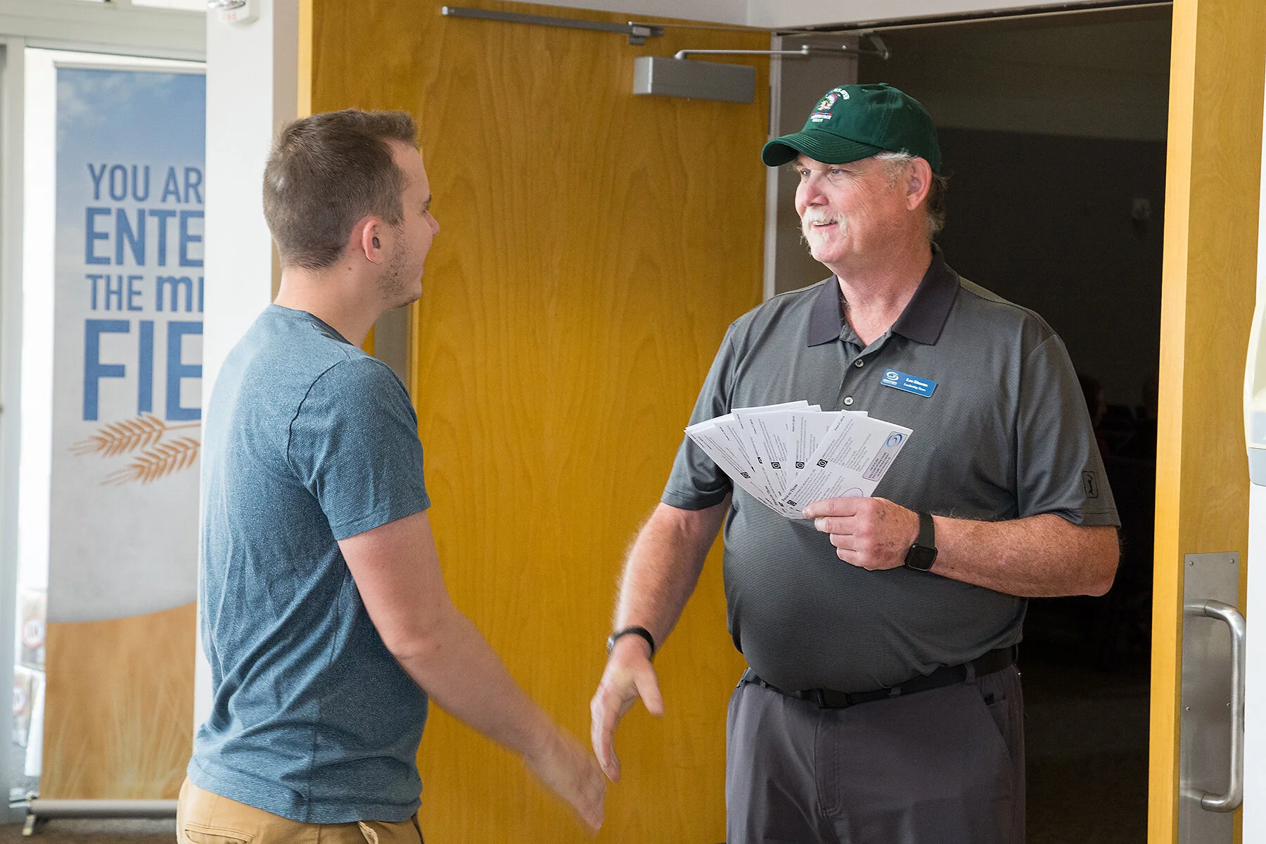 A young man shaking hands with an older man at the entrance of a building. The older man is wearing a dark gray polo shirt, a green cap, and has a name tag. The young man is wearing a gray T-shirt and khaki shorts. The older man is holding pamphlets 