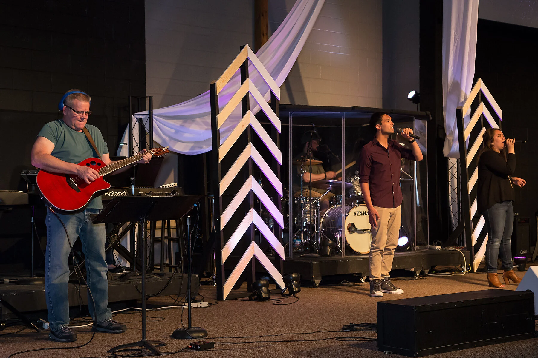 Musicians performing on stage with a guitarist, drummer, and two singers under stage lighting and decorative wooden panels.