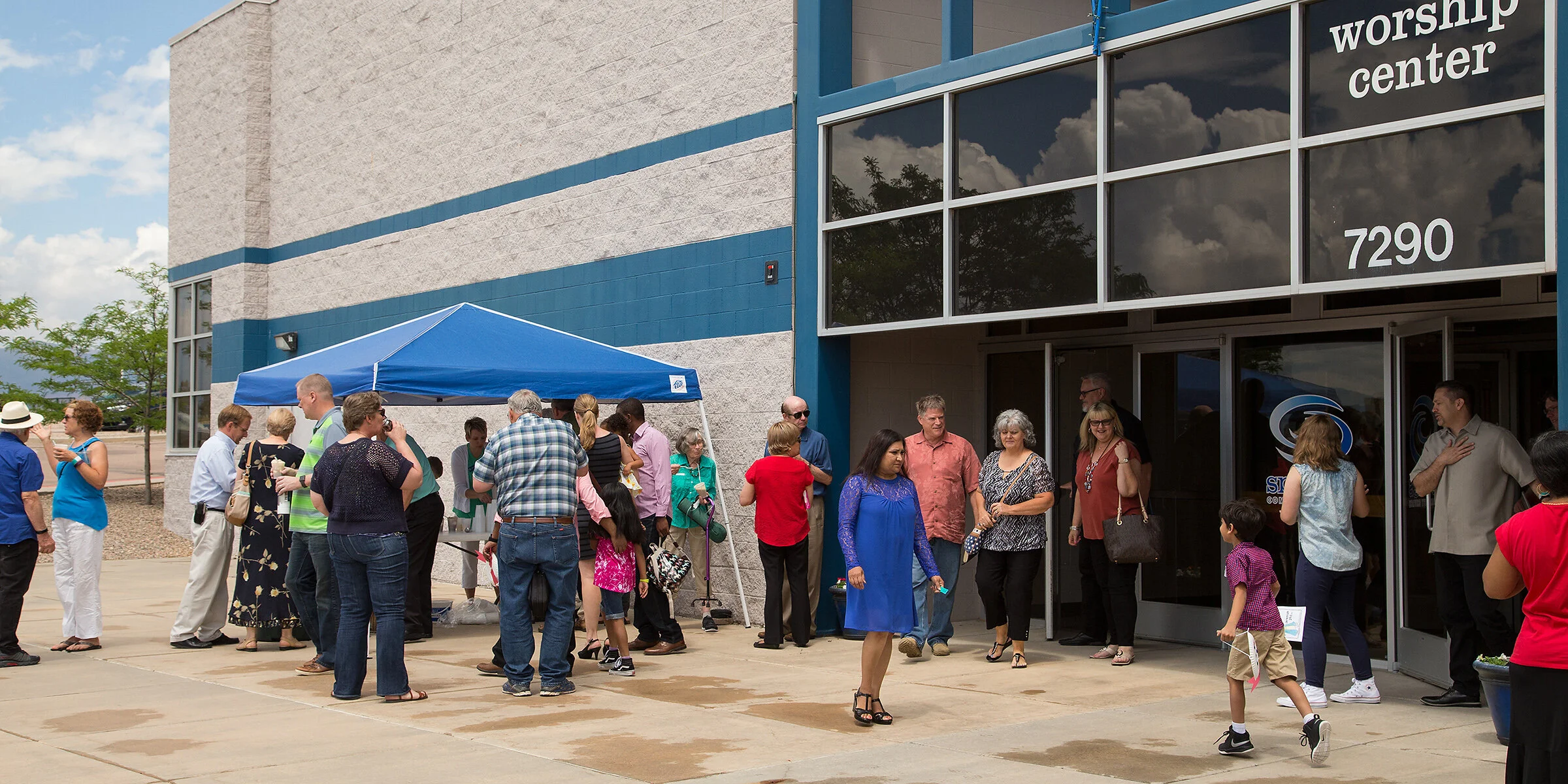 A group of people gathered outside a worship center building with a blue awning and glass doors, engaging in conversation.