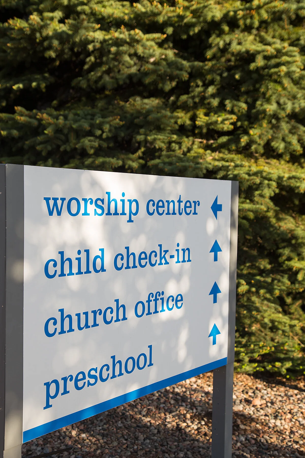 Signboard outdoors with directions for worship center, child check-in, church office, and preschool, all pointing straight ahead, against a backdrop of green pine trees.