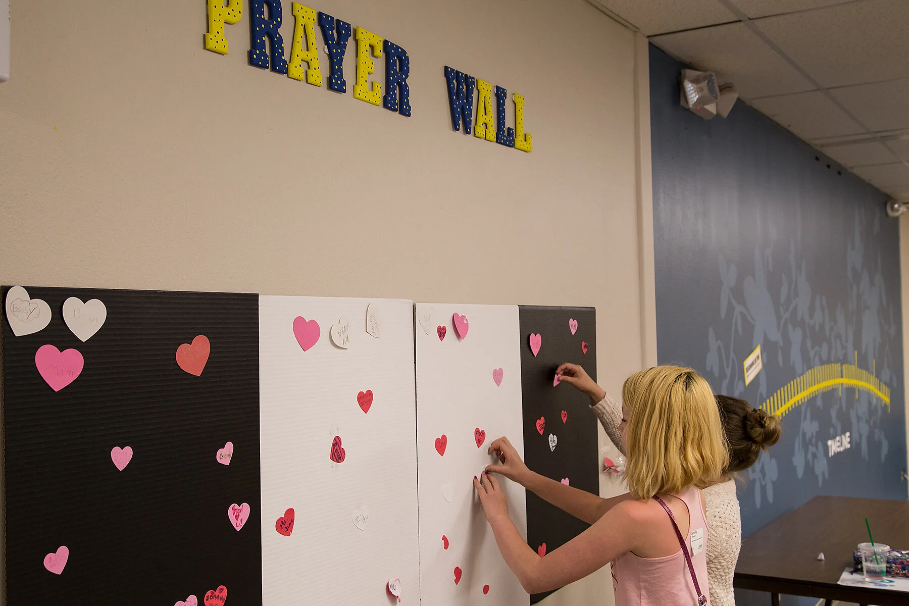 Two women are decorating a wall with pink and red heart-shaped paper cutouts for a celebration. The wall has a sign that reads 'Prayer Wall' in colorful, marquee-style letters. One woman with blond hair is attaching a heart to the wall, and the other