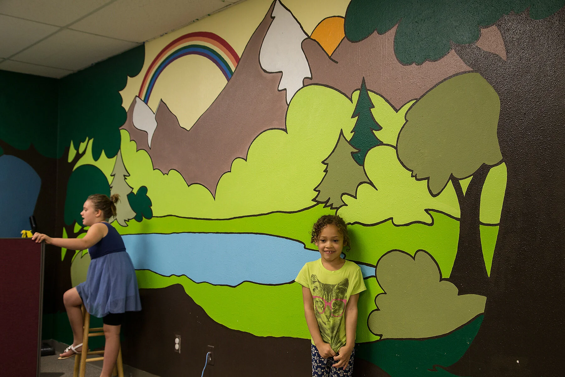 Two young girls are standing in front of a colorful mural of a mountain landscape with trees, a river, a rainbow, and a sun.