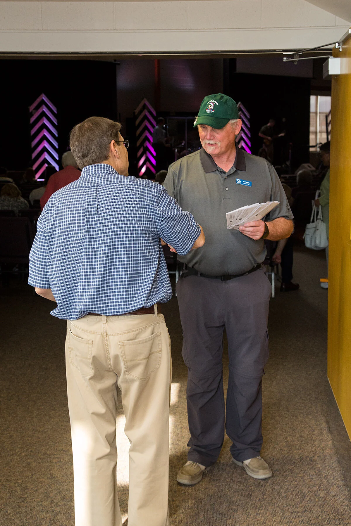 Two men are having a conversation indoors at a public event, with a dark stage and purple lighting in the background.