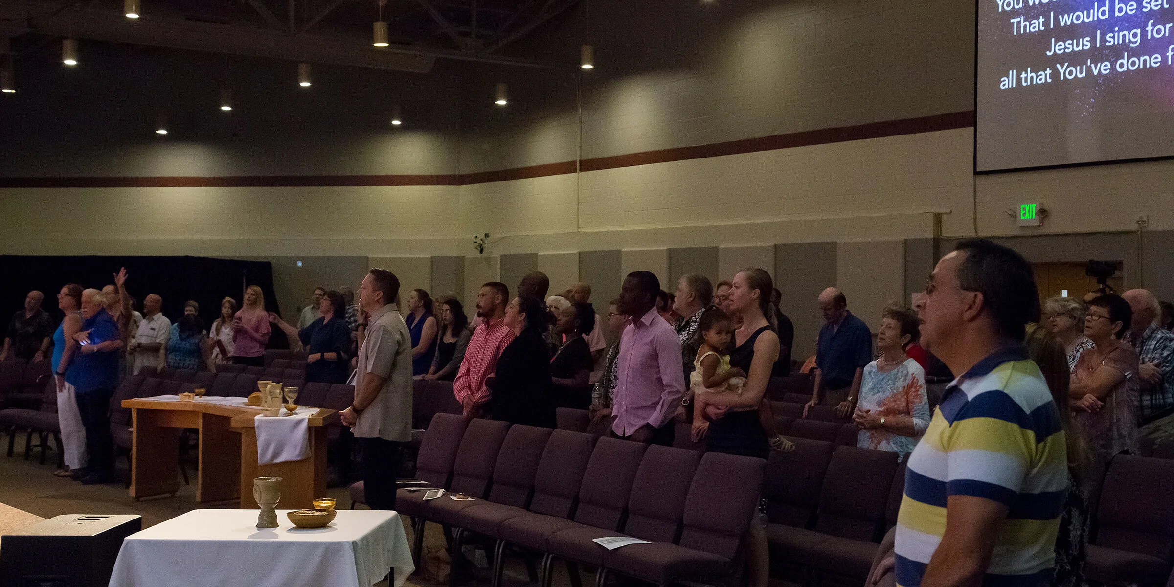 People standing in a church or auditorium, some with eyes closed or hands raised, participating in a religious service or worship. There are tables with bread and cups in the foreground and a large screen displaying song lyrics on the right.