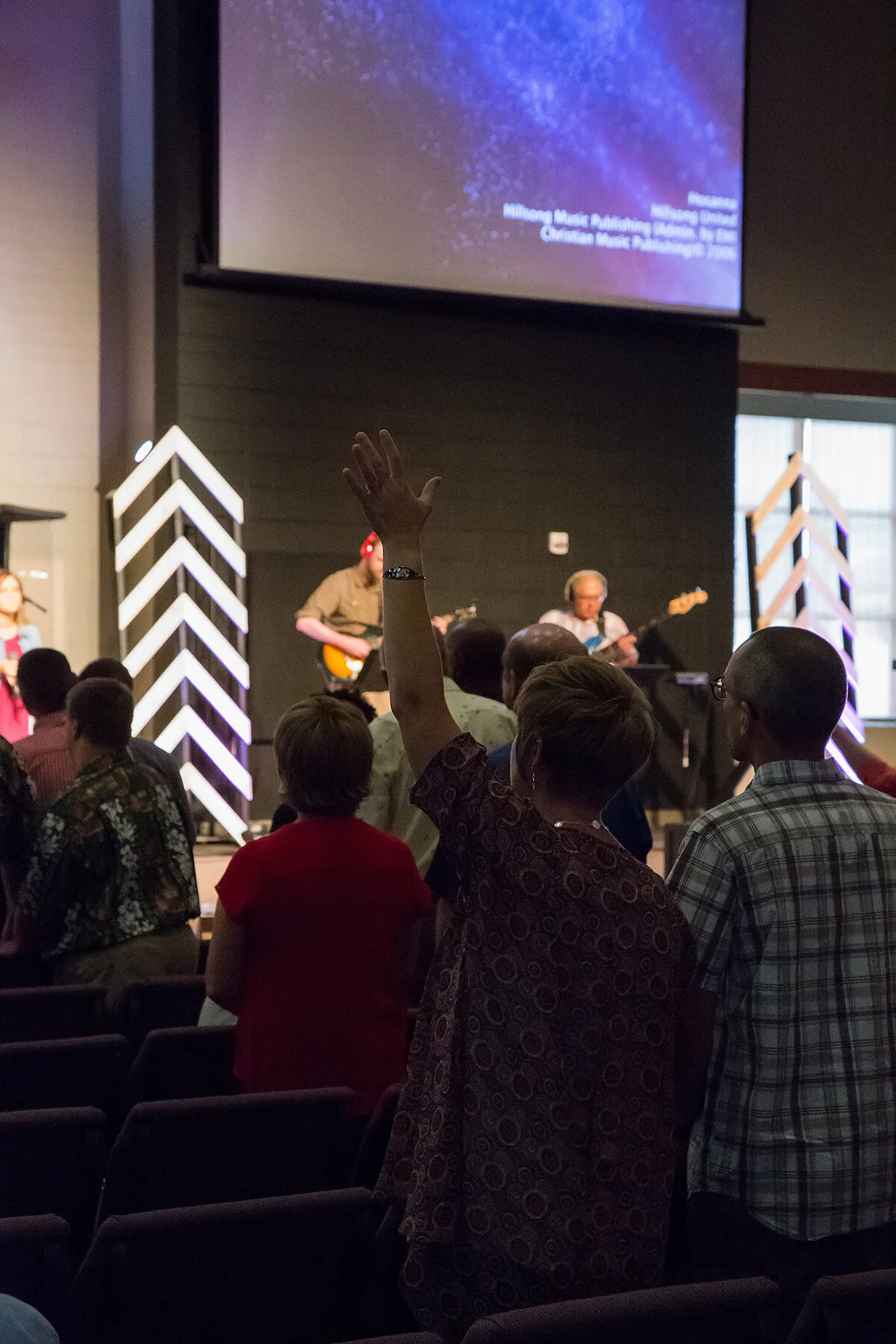 People attending a church service or event with a band playing on stage, one person raising their hand in the audience, and a large screen displaying song lyrics or presentation behind the stage.