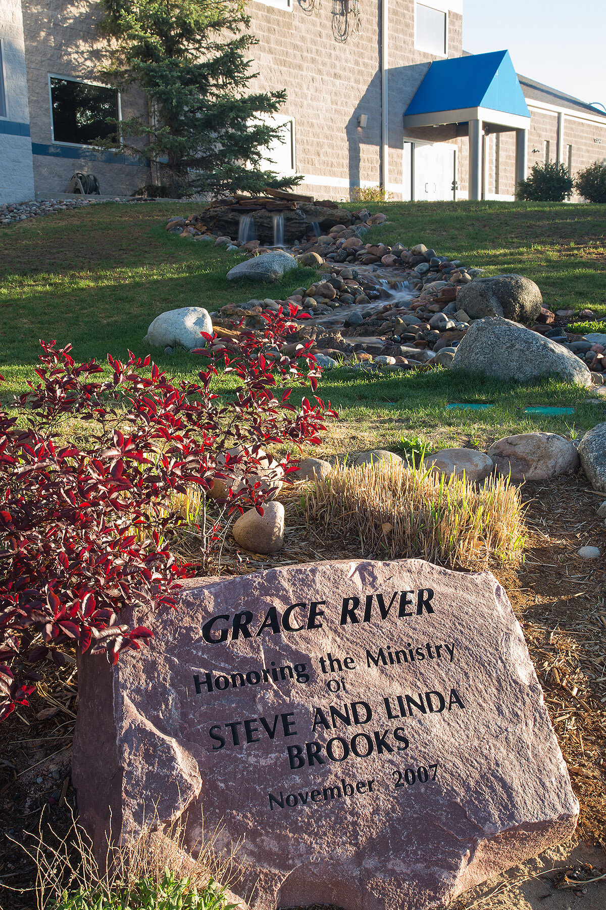 A memorial stone in the foreground with the inscription 'GRACE RIVER Honoring the Ministry of STEVE AND LINDA BROOKS November 2007,' surrounded by plants and rocks. In the background, there is a landscaped yard with grass, a small rock waterfall, and
