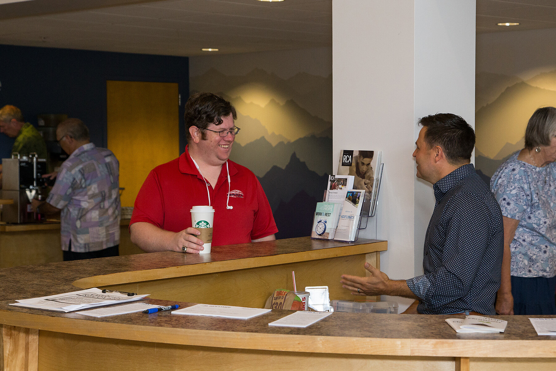 A man in a red polo shirt at a reception desk talking to another man in a dark, patterned shirt. The man in red is holding a Starbucks coffee cup and smiling. In the background, there are other people and a wall with a mountain mural.