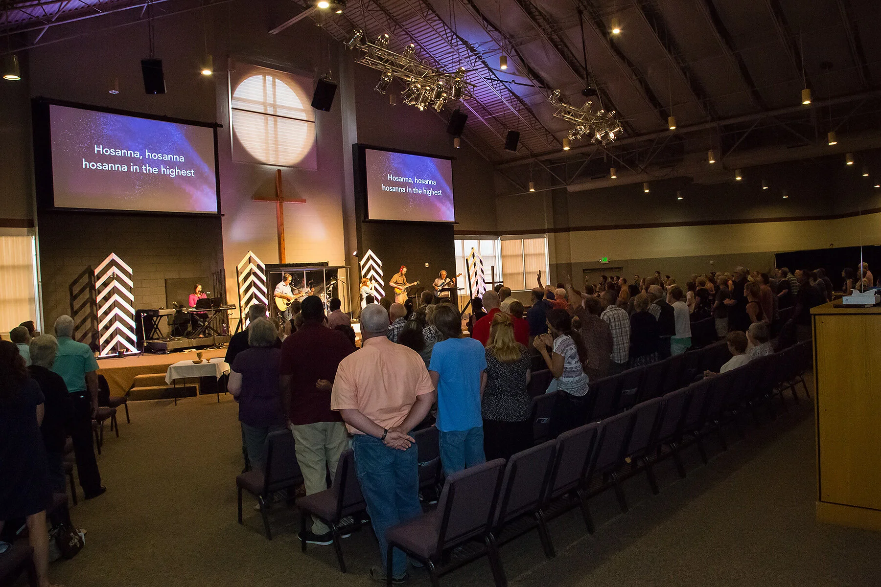 A church service with a congregation standing and singing, led by a band on stage with a large cross in the background and lyrics displayed on two screens.