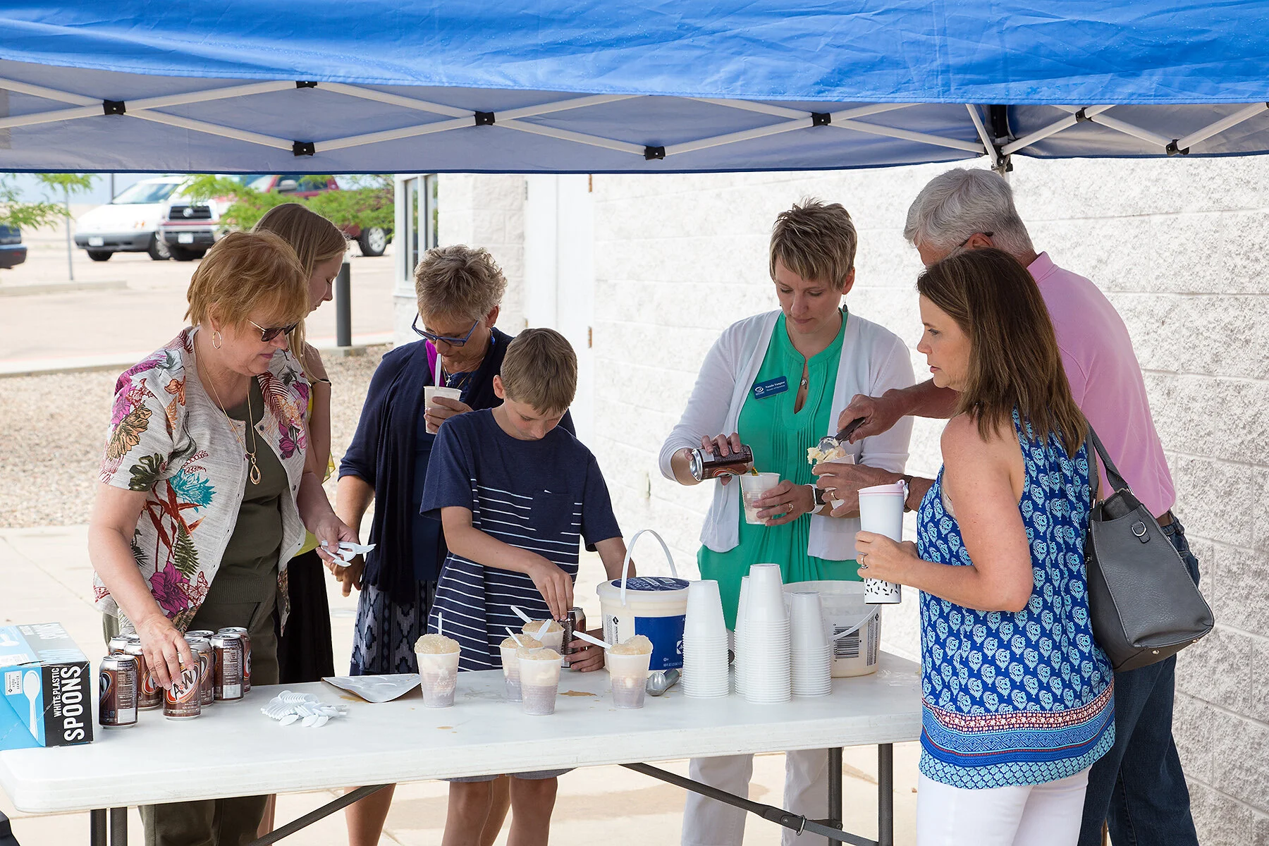 People gathered around a table under a blue canopy, serving and eating ice cream cones outdoors.