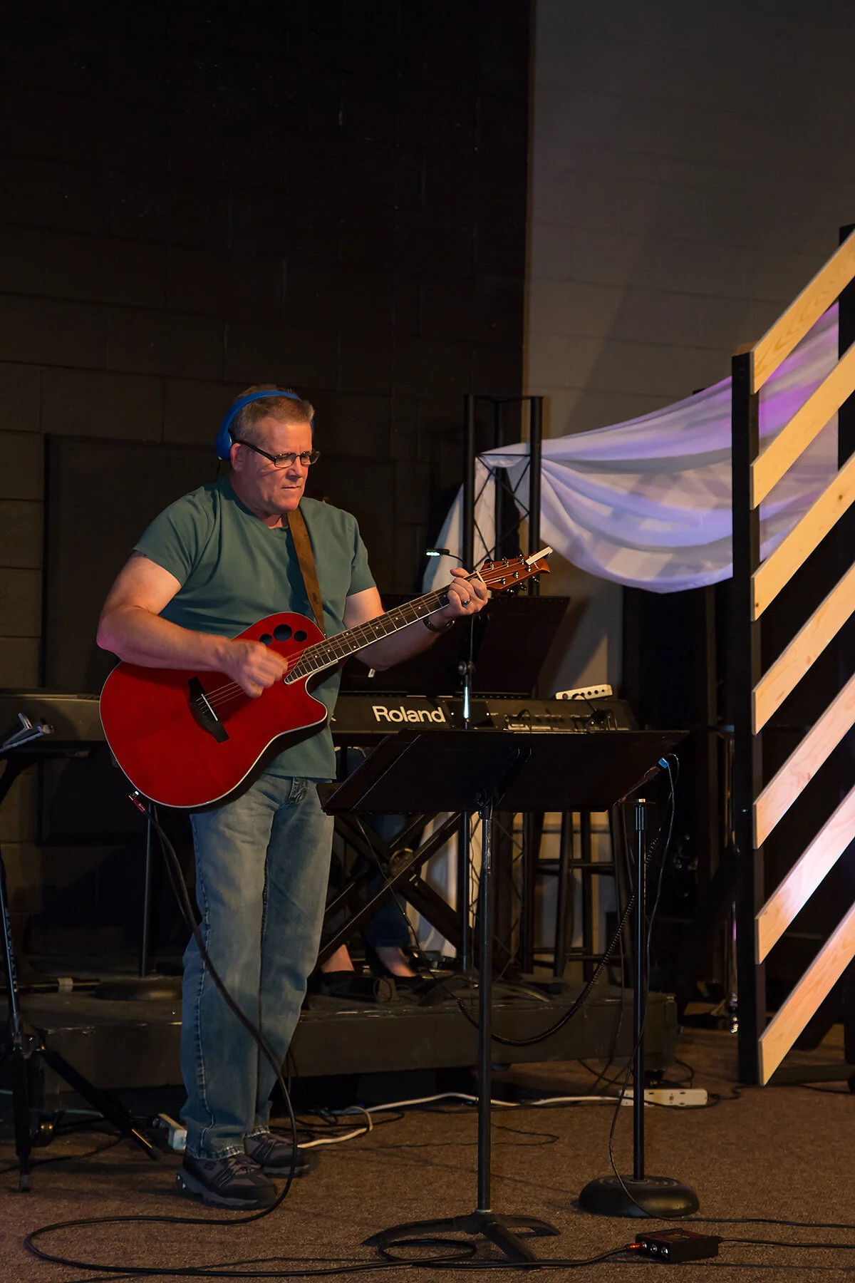 A man wearing glasses and headphones plays an acoustic guitar on stage with a music stand, keyboard, and digital equipment in background.